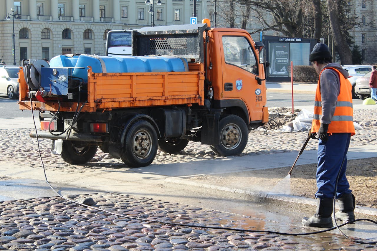 A city worker cleans a cobblestone street with a hose and truck in an urban area.